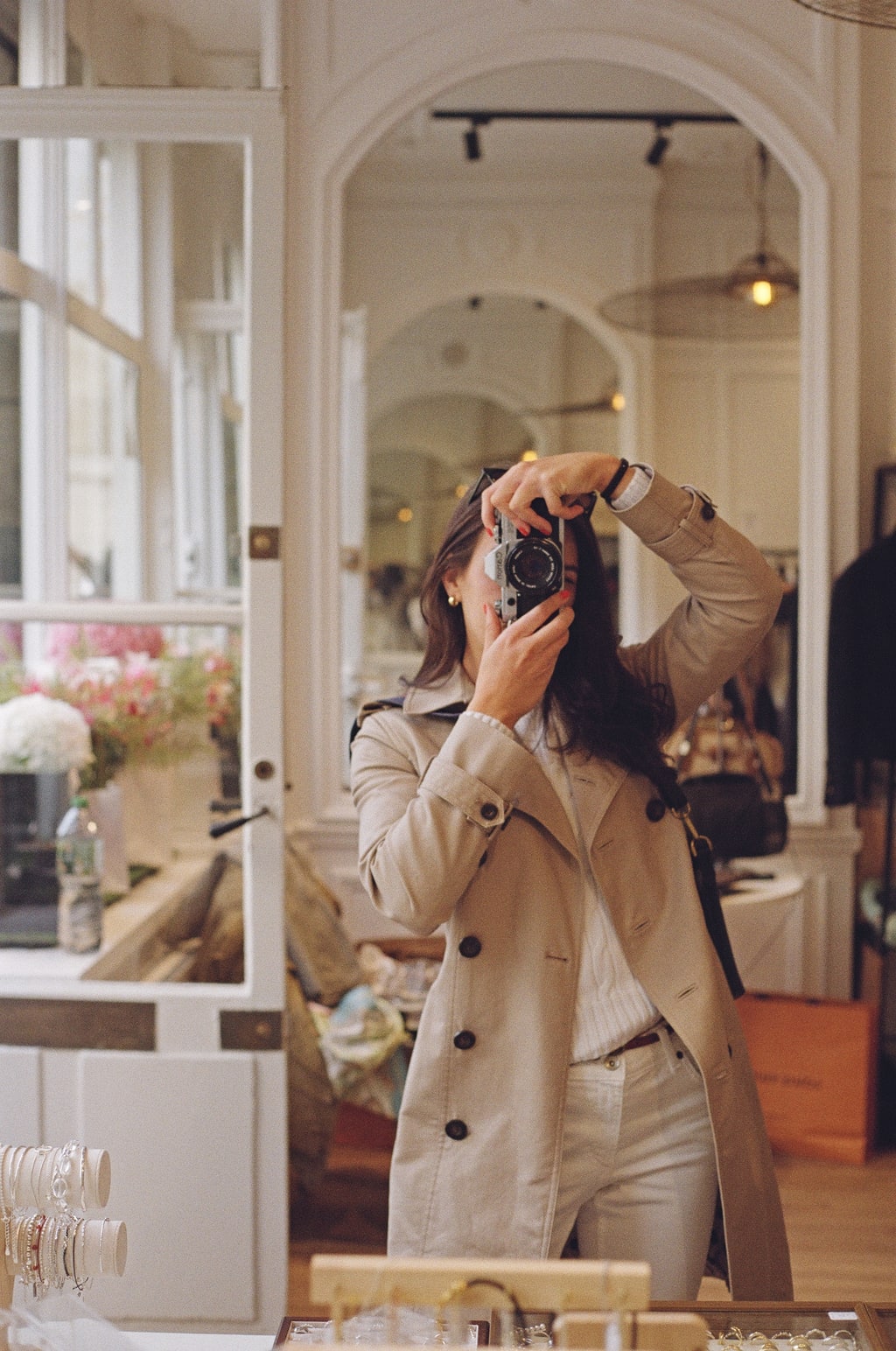 Alex Cros photographed in a mirror, holding a 35mm rangefinder camera to her eye in a bright shop interior.