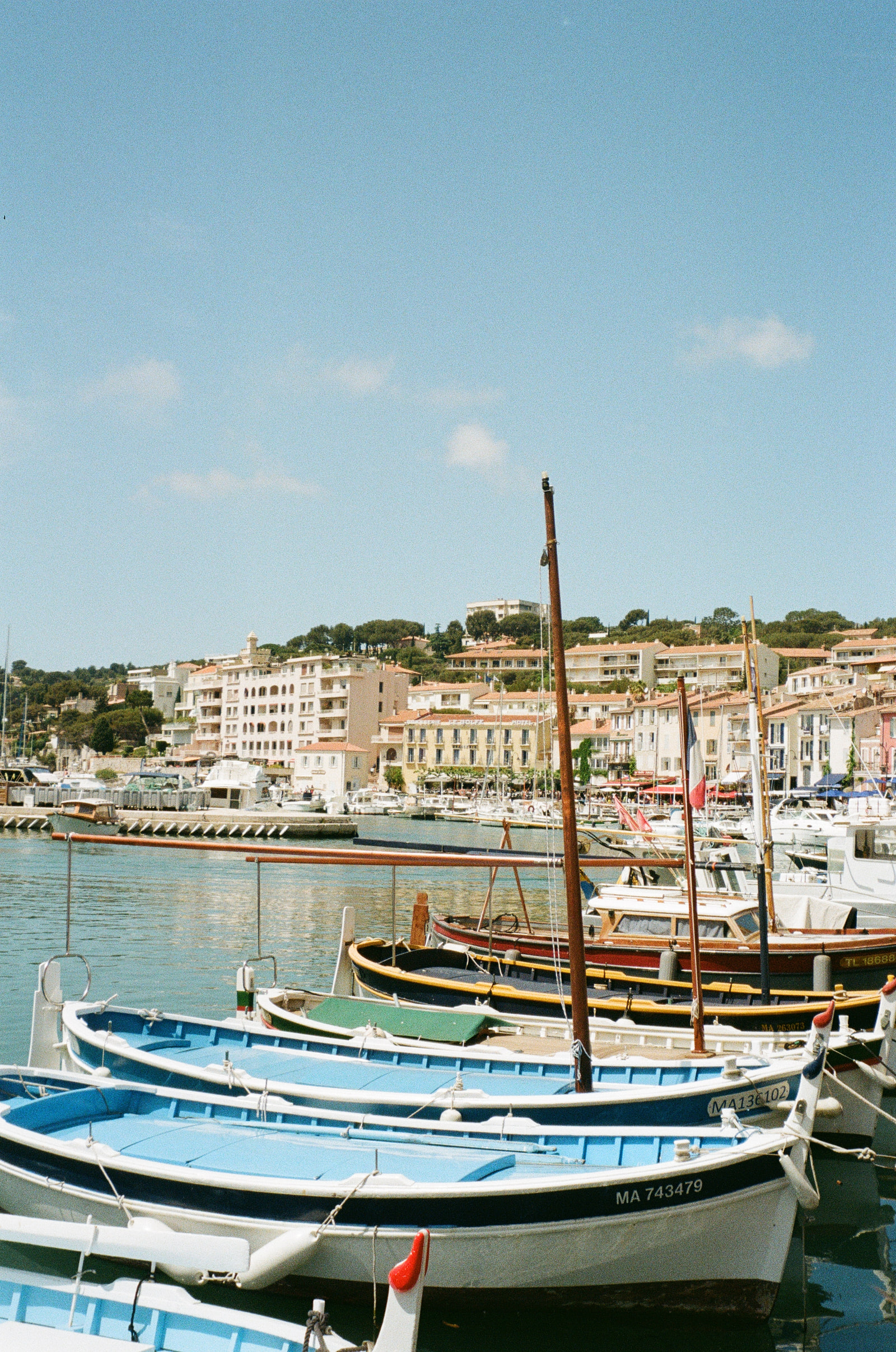 Traditional wooden barquettes moored in the harbour at Cassis, painted pale blue, green and yellow, with the terracotta roofs of the town and a wooded hill rising behind them.
