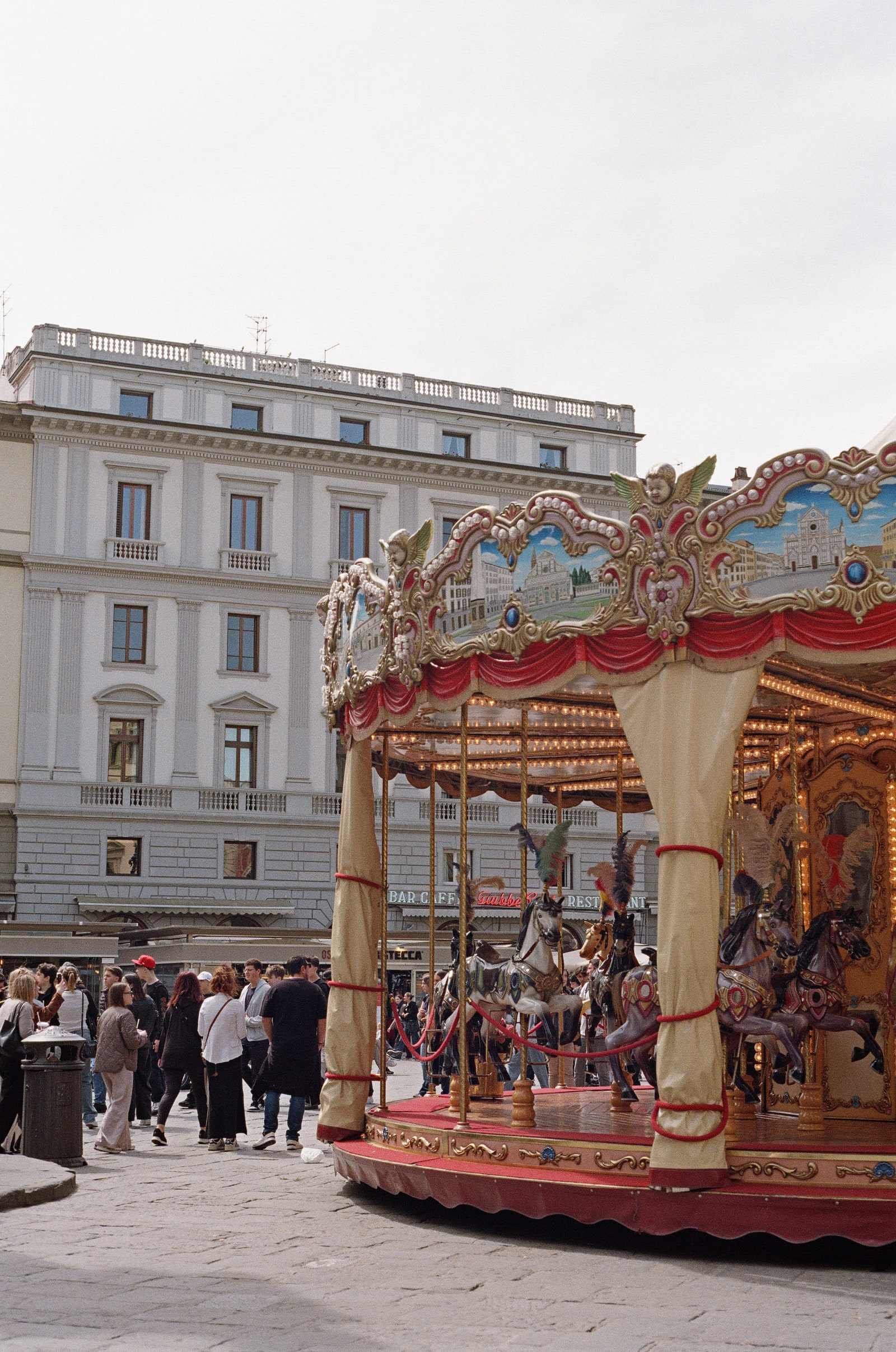 A gilded carousel standing in Piazza della Repubblica in Florence, its canopy painted with views of the city, horses waiting under strings of bulbs, and a pale palazzo facade behind it with a crowd along the edge.