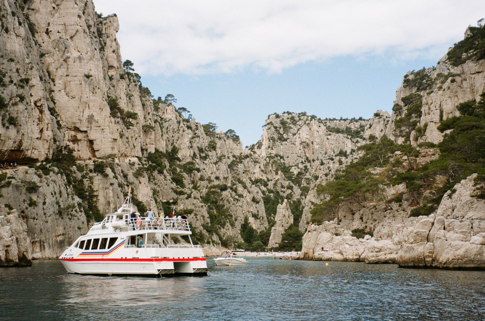 A white tour catamaran with a red stripe moored in a calanque near Cassis, passengers standing at the rail looking up at the limestone cliffs, pines along the rock, and a small beach at the head of the inlet.