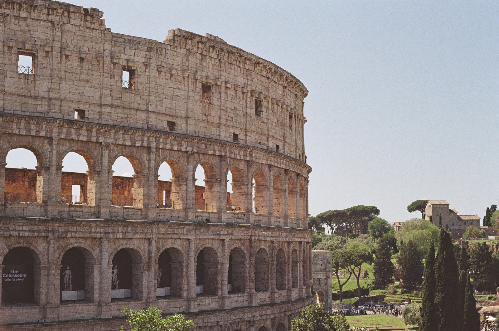 The outer wall of the Colosseum in Rome photographed from above in flat afternoon light, its travertine arches stacked in three tiers against a pale hazy sky, with umbrella pines on the ridge beyond.