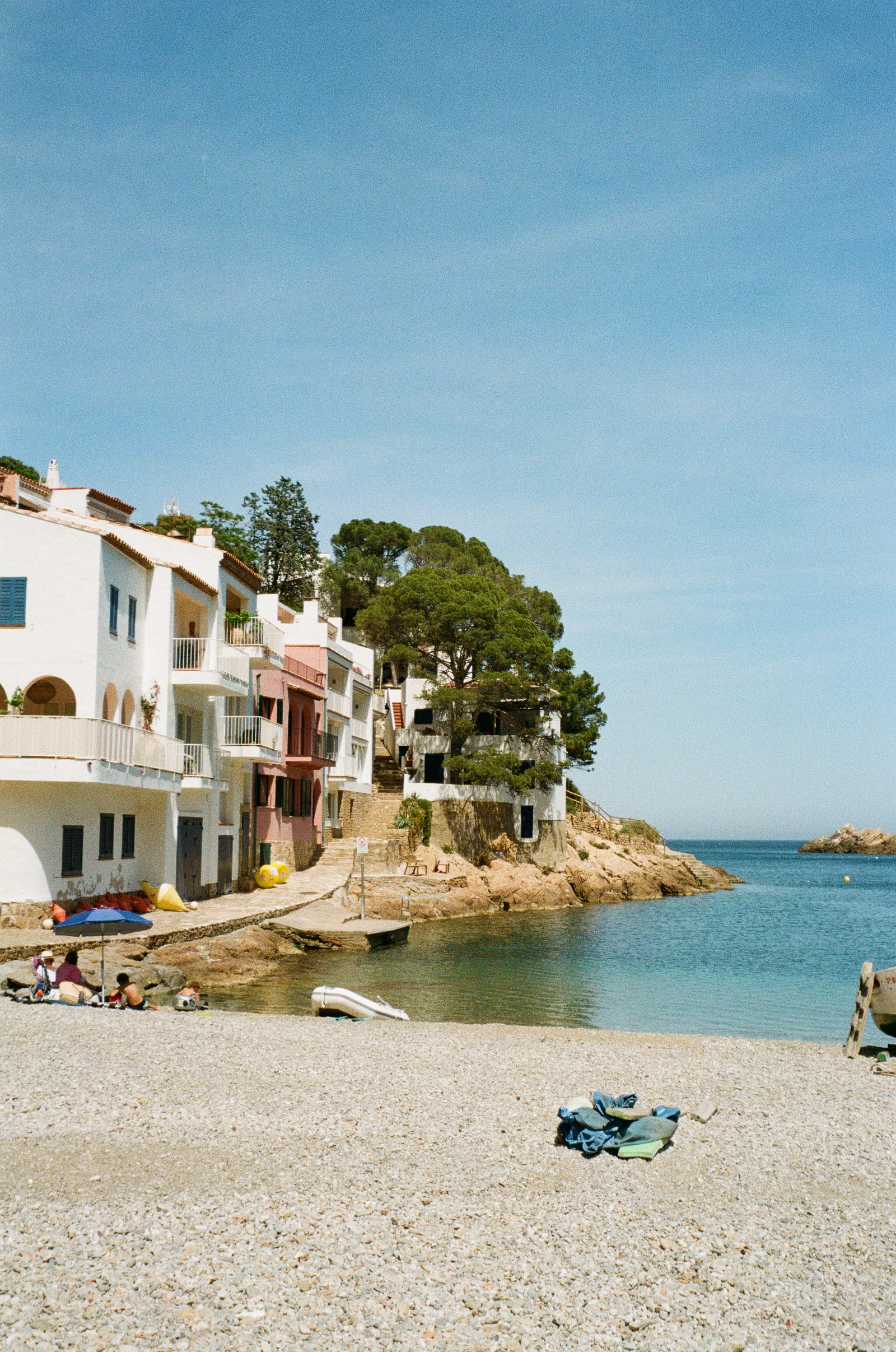 A small cove at Begur on the Costa Brava at midday, white and pink houses stepping down to the water beside a stone pine, a pebble beach in the foreground and clear turquoise water reaching out to a rock offshore.
