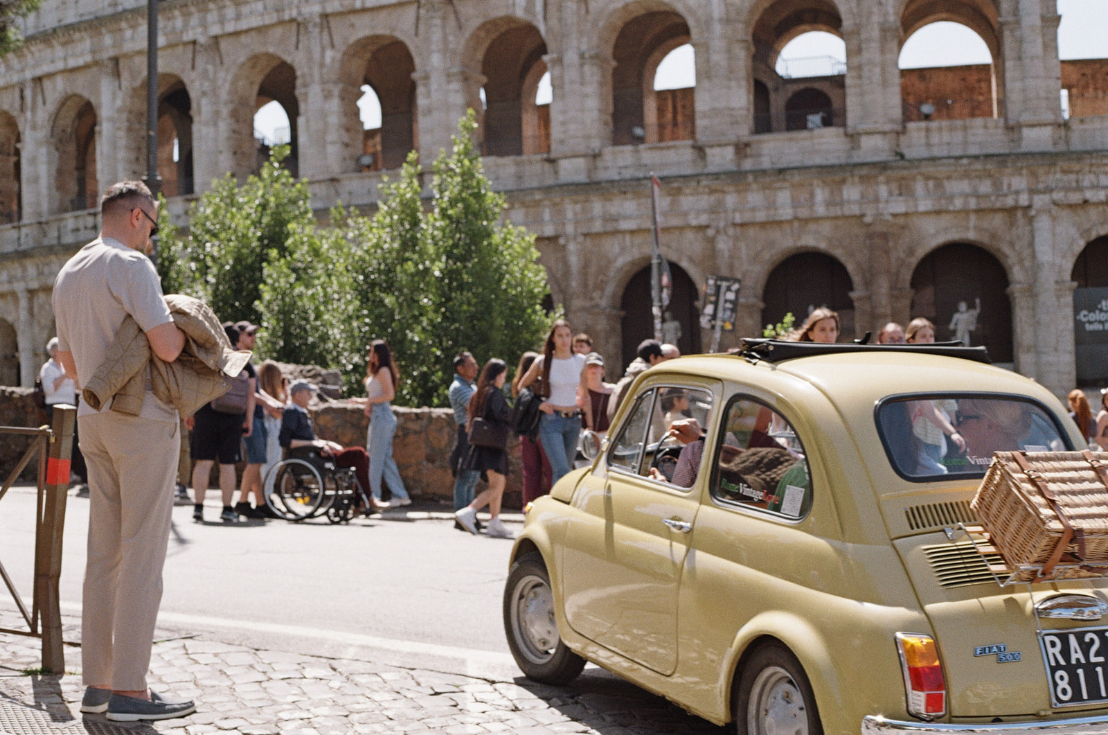 A cream-yellow vintage Fiat 500 with a wicker picnic basket strapped to its rear rack, passing in front of the Colosseum in Rome, a man in a linen shirt watching from the pavement as tourists cross behind.