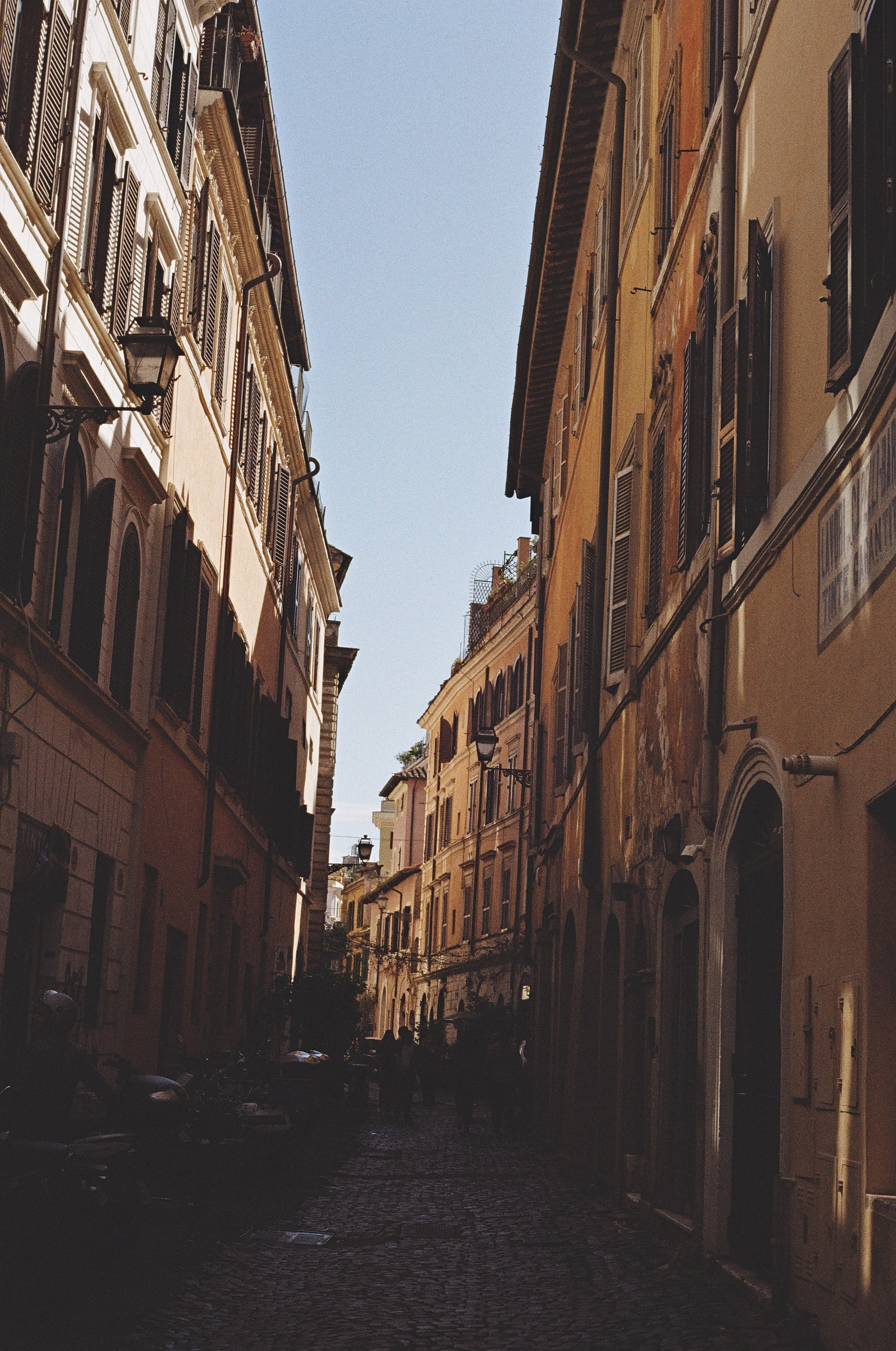 A narrow cobbled street in Florence falling into deep shadow, wrought-iron lamps projecting from ochre facades on both sides, and a wedge of pale sky between the rooflines above.