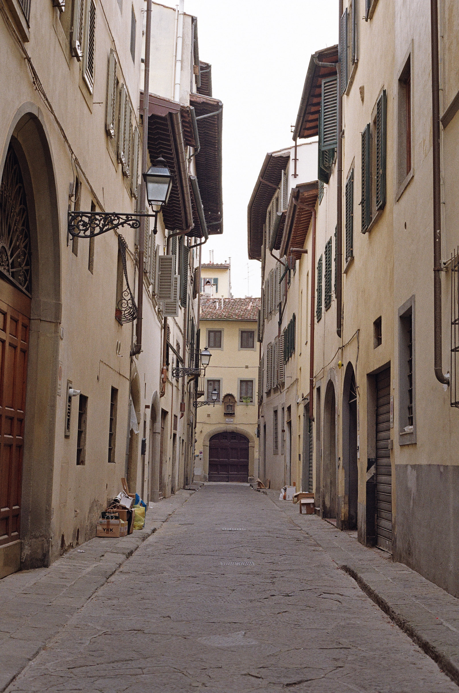 An empty stone lane in Florence with shuttered buildings on both sides, flattened cardboard boxes stacked against the kerbs, iron lamps overhead, and a dark arched gate closing the far end.