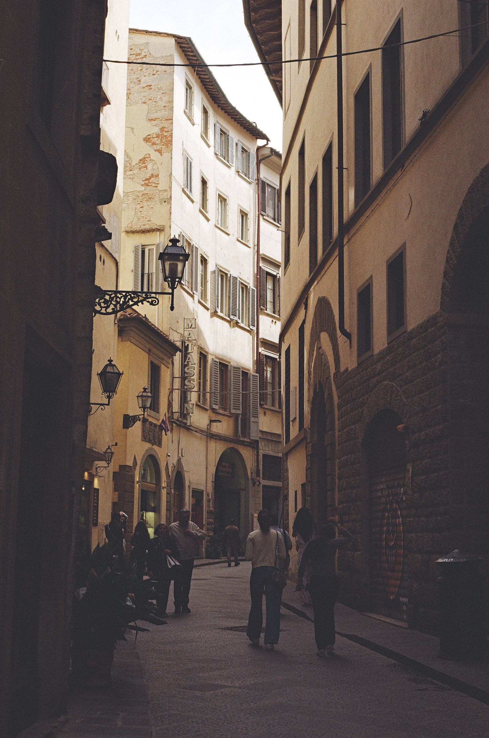 A narrow Florence street with a row of iron lanterns projecting one behind another from the left-hand wall, figures walking away into the distance, and a sunlit facade closing the view at the end.