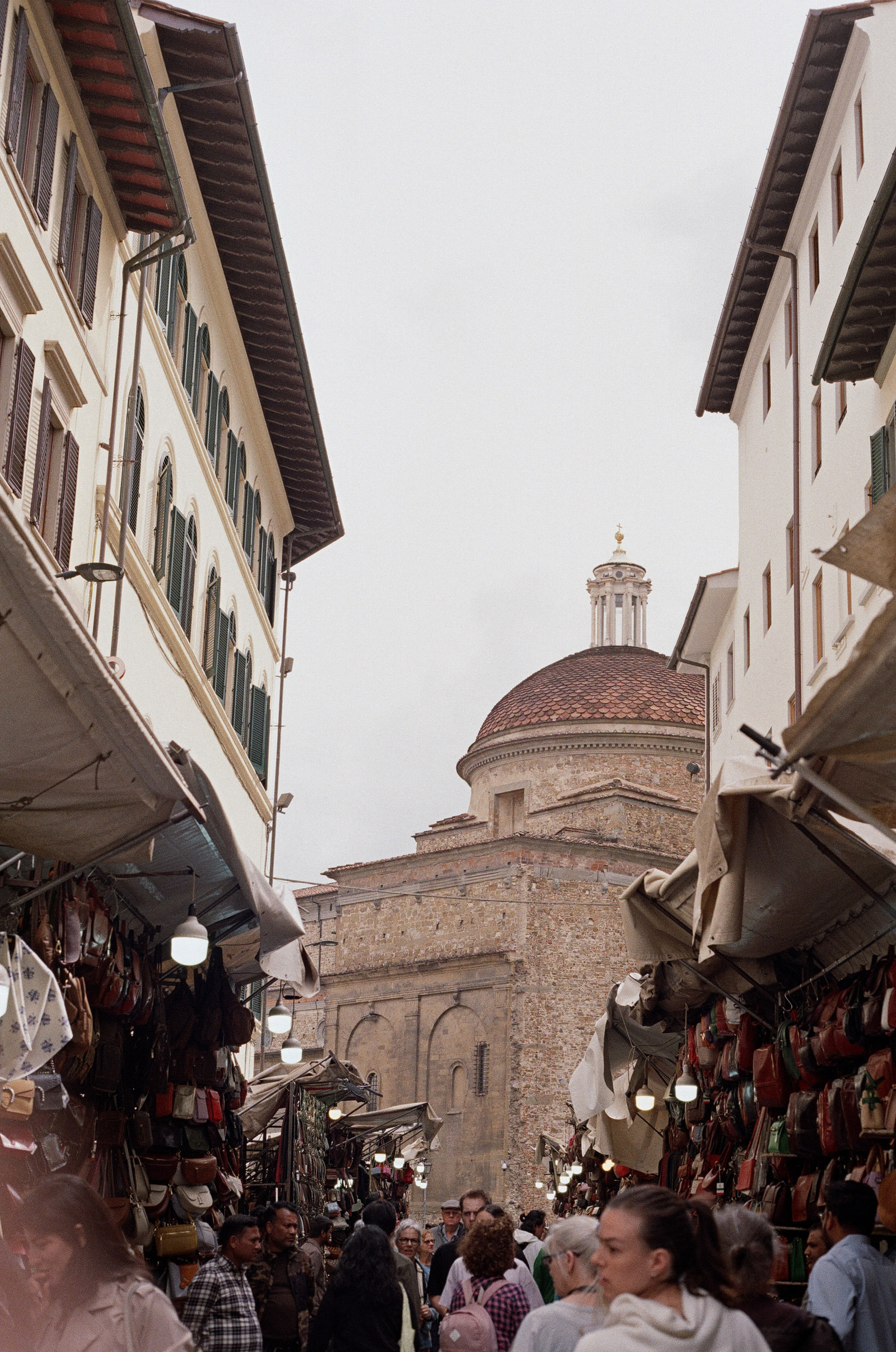 Leather stalls crowding both sides of a street at the San Lorenzo market in Florence, bare bulbs hanging above the bags and belts, and the tiled dome and lantern of San Lorenzo rising in the gap beyond.