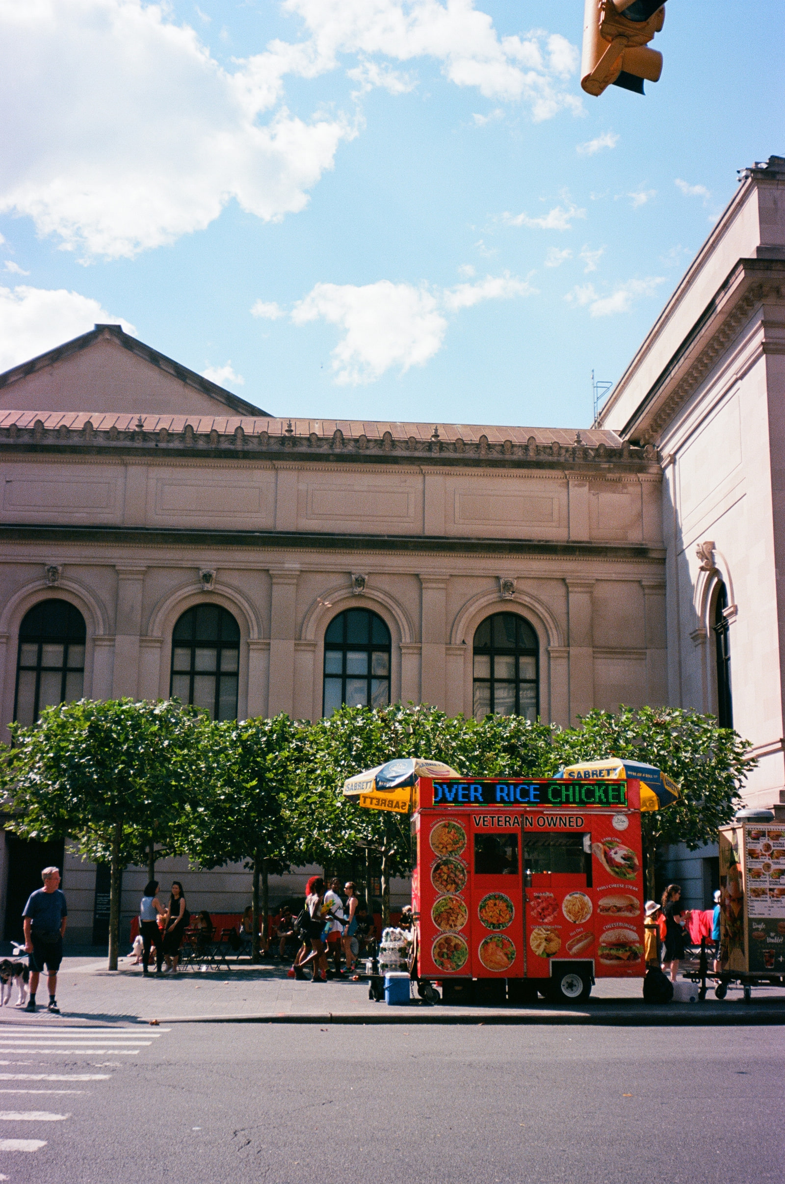 A red halal food cart with striped Sabrett umbrellas parked under pollarded trees outside the Metropolitan Museum of Art on Fifth Avenue, with a man walking a dog across the empty street in the foreground.