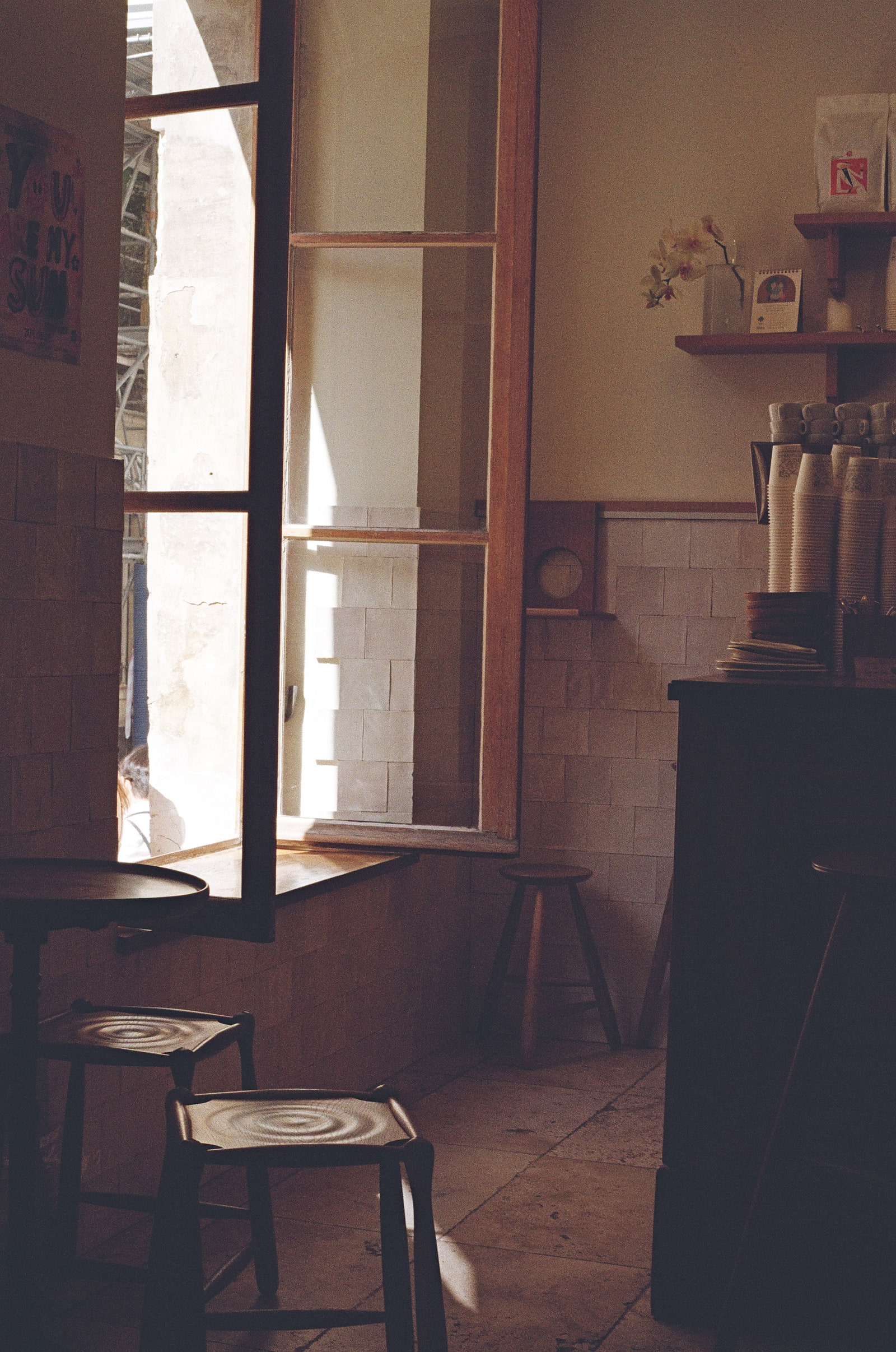 The inside of a quiet Paris cafe, an open window throwing light across a white tiled wall, wooden stools and a small round table in shadow, and stacked paper cups on the counter to the right.