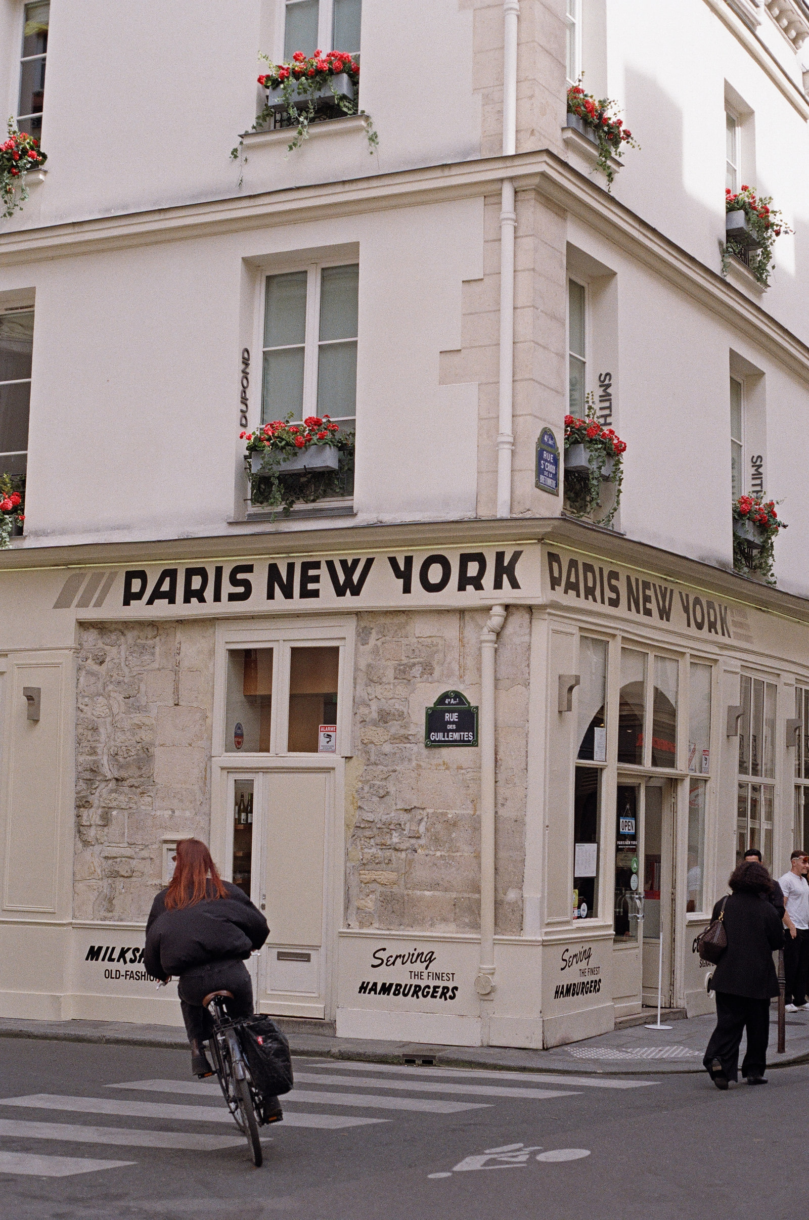A cream corner building in the Marais lettered PARIS NEW YORK in bold black type, red geraniums in the window boxes above, and a red-haired cyclist crossing the street in front of it.