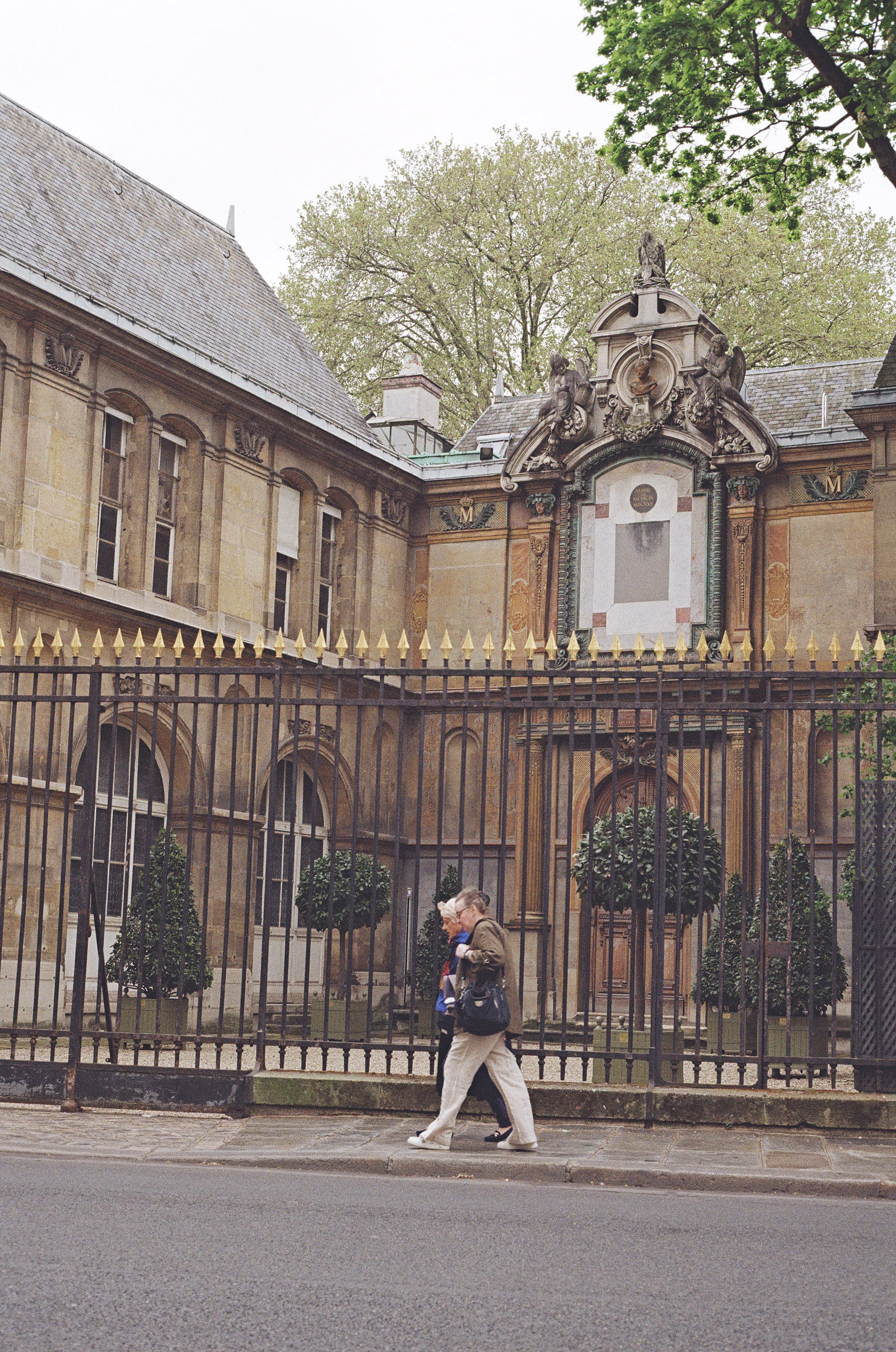 An elderly couple walking past a tall iron railing in Paris, the gilded spear tips of the railing running across the frame, and an ornate carved portal with clipped bay trees behind them.