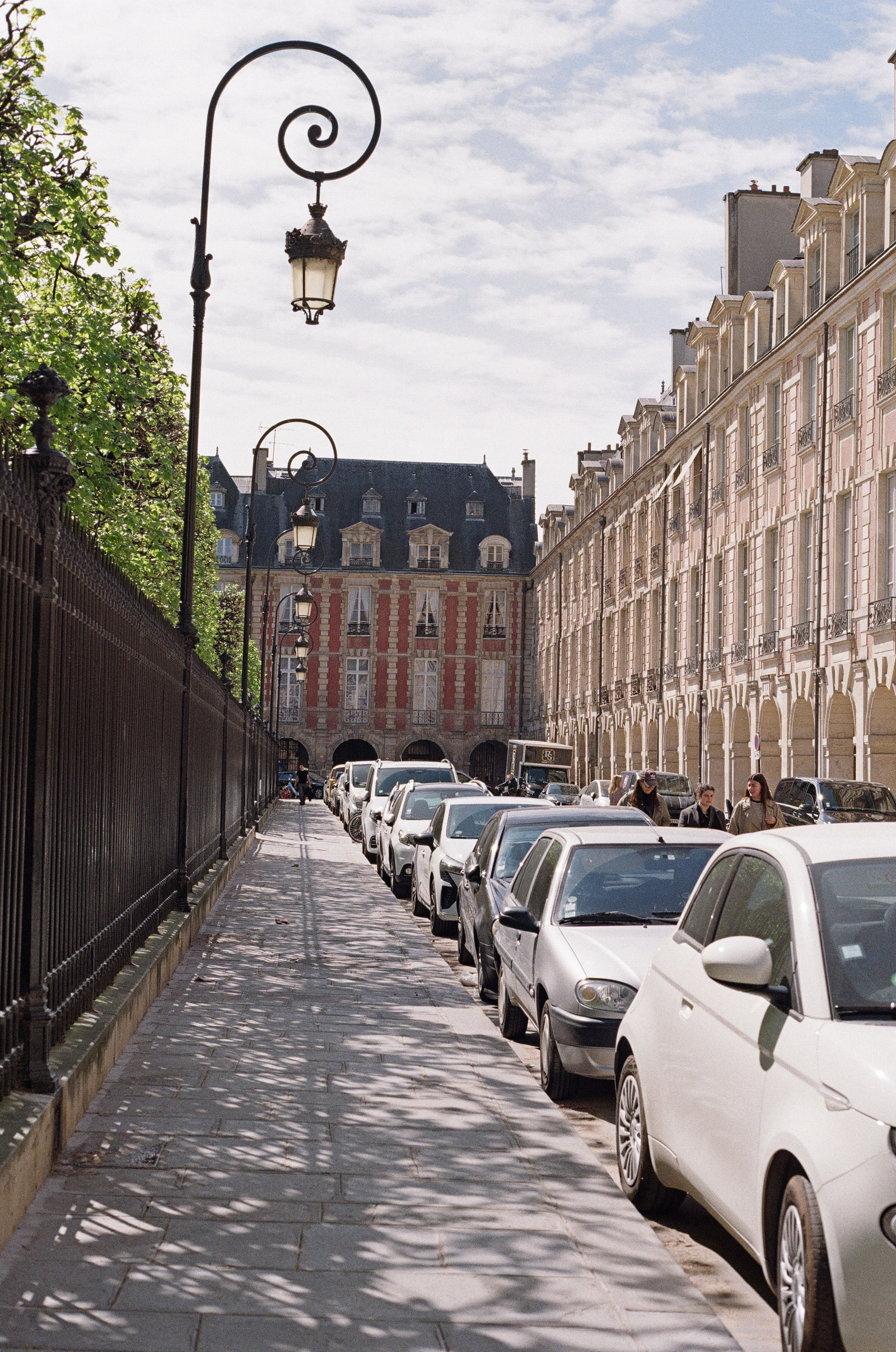 A pavement running along Place des Vosges in Paris with three scrolled iron lamp standards receding into the distance, cars parked along the right, and the brick and stone Pavillon de la Reine closing the view.