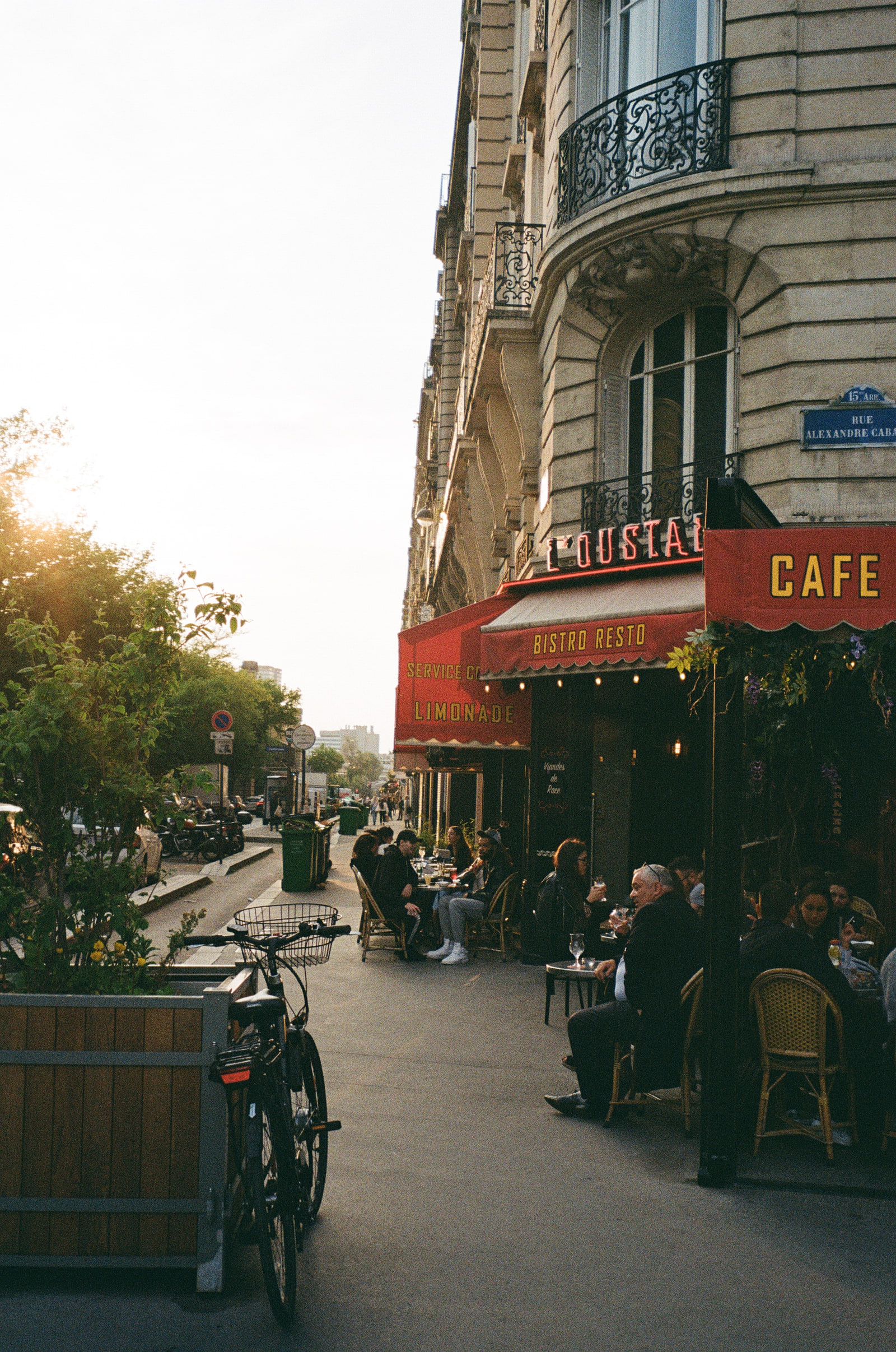 A Paris café on a corner in the fifteenth arrondissement at golden hour, its red awning lettered Bistro Resto and Limonade, people seated at rattan chairs on the pavement and low sun flaring through the trees opposite.