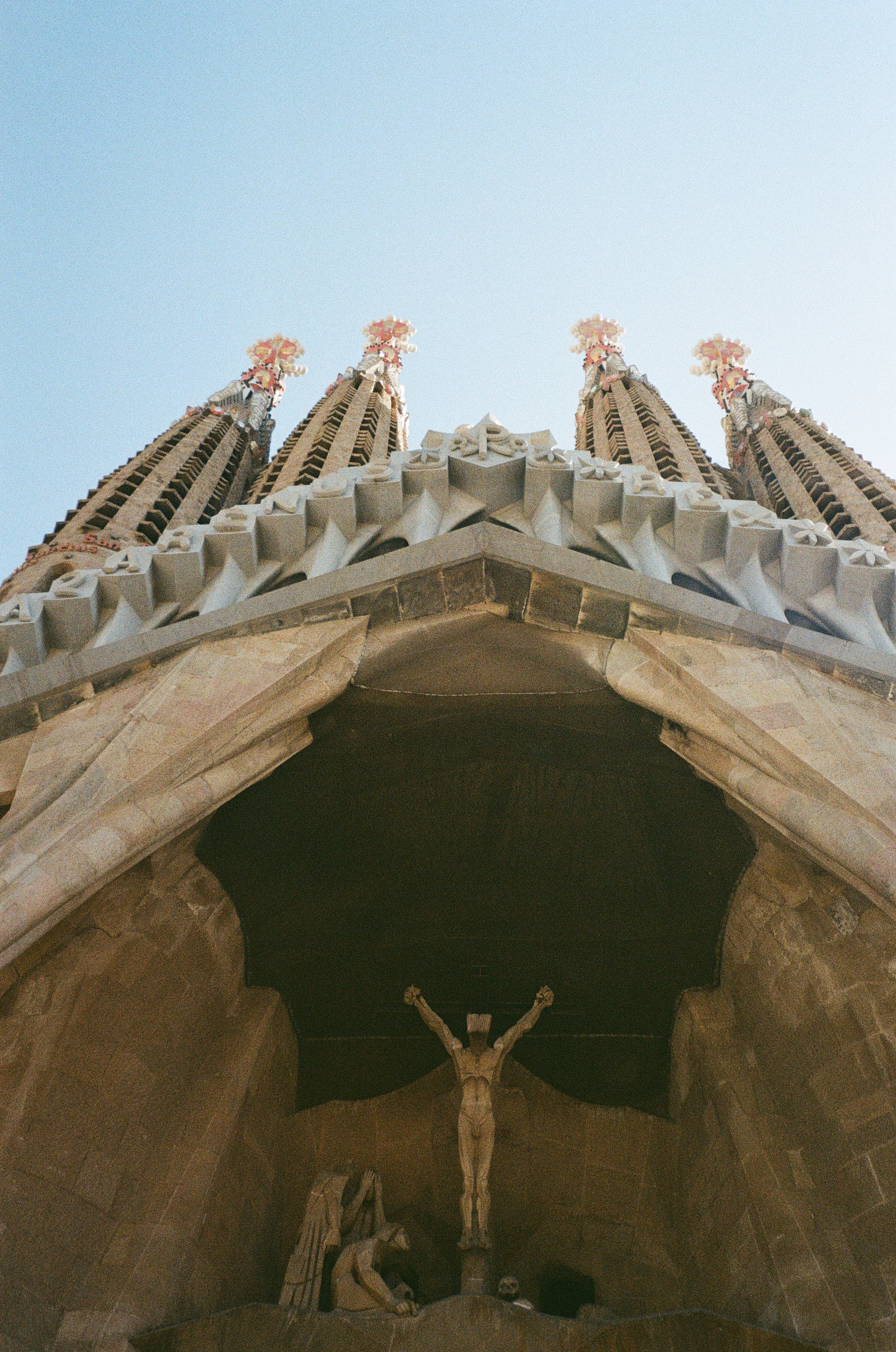 The Sagrada Família in Barcelona photographed from directly below, four spires converging against a pale sky above a sharply angled gable, with the carved crucifixion group of the Passion facade set deep in the shadowed portal beneath.