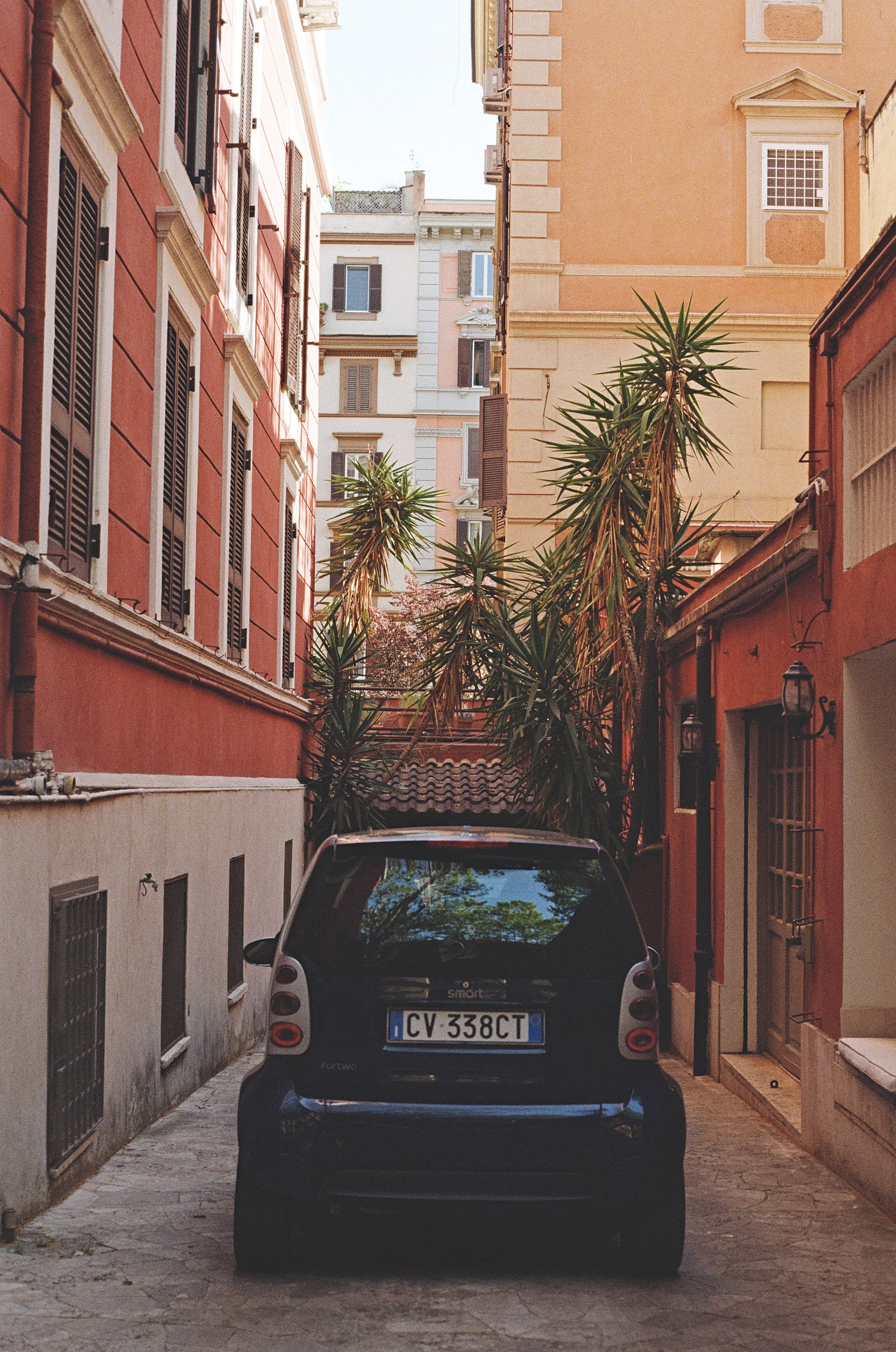 A black Smart fortwo parked in a narrow Roman alley barely wider than the car, red and terracotta walls rising on both sides, and overgrown yuccas leaning across the passage above it.