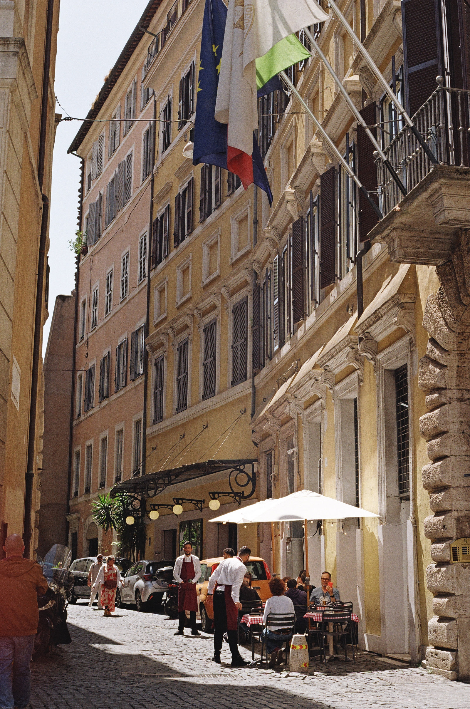 European, French, and Italian flags hanging into the frame above a Roman street, two waiters in burgundy aprons standing beside red-checked tables under a white umbrella on the cobbles below.
