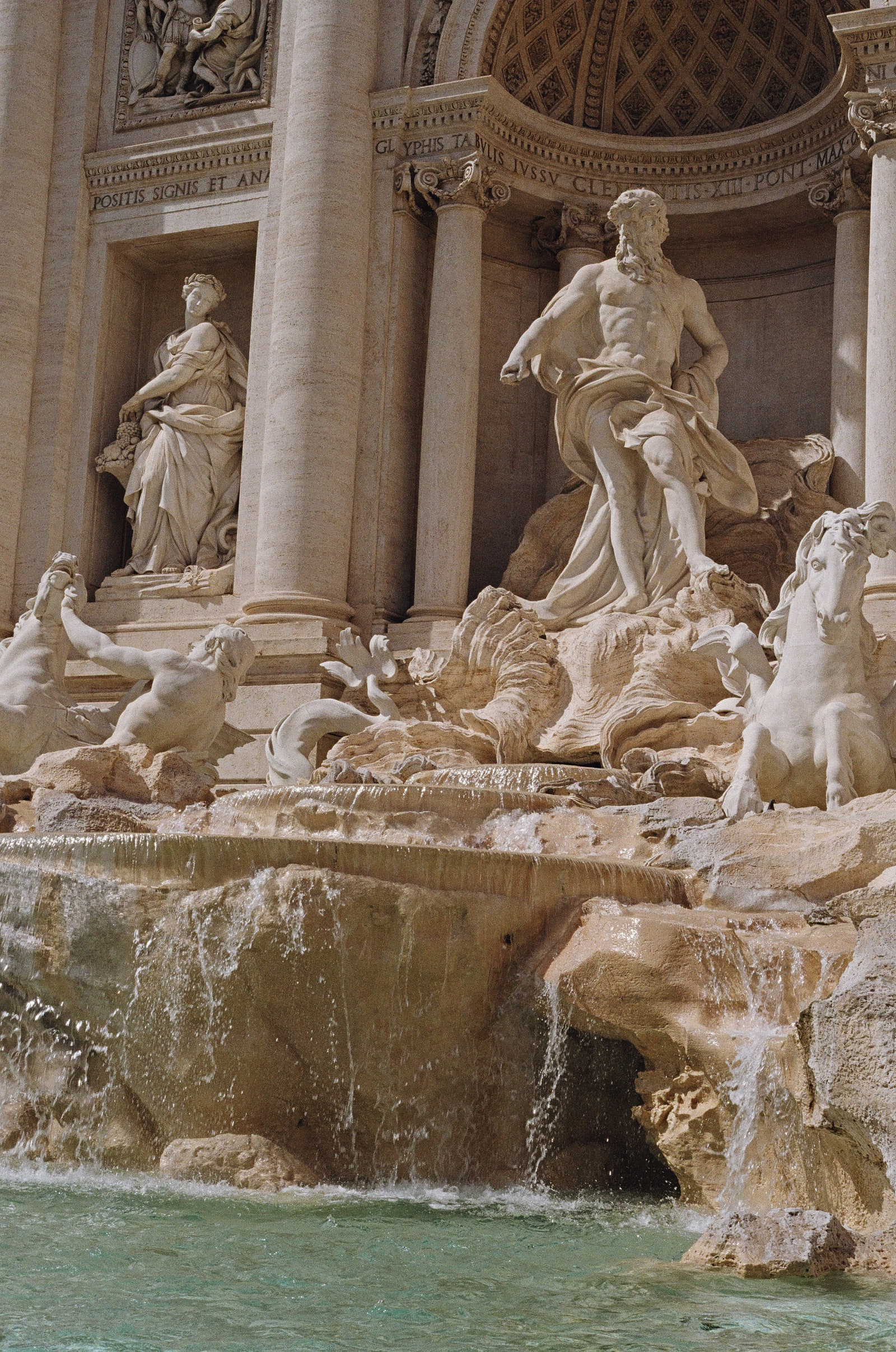 A close view of the Trevi Fountain in Rome, Oceanus and his sea horses carved in white marble above rough travertine rock, with jade-green water pouring across the foreground.