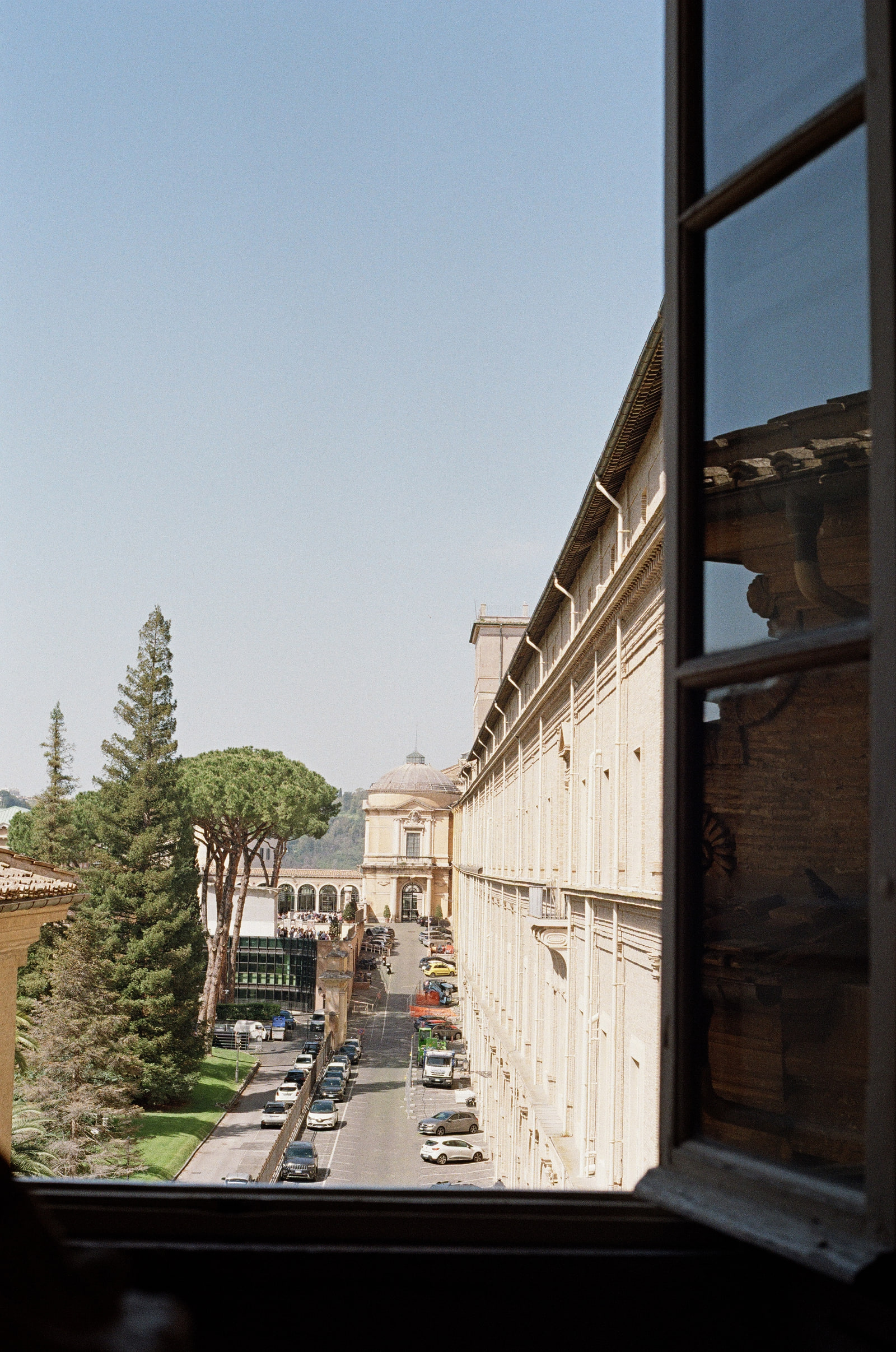 A view through an open museum window in the Vatican, the dark window frame filling the right of the picture, umbrella pines and a domed pavilion beyond, and cars parked along the wall below.