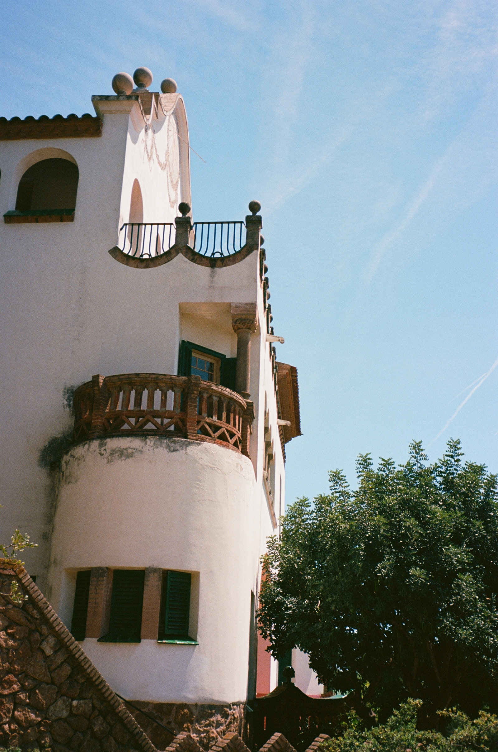 A whitewashed Catalan villa seen from below against a deep blue sky, with a curved brick balcony, dark green shutters, stone finials along the roofline and a contrail crossing overhead.